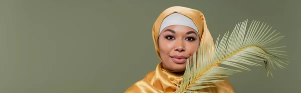Giovane donna multirazziale in hijab di seta gialla sorridente alla macchina fotografica vicino a foglia di pianta tropicale isolata su verde, striscione — Foto stock