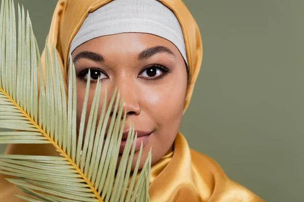 Retrato de mujer musulmana multirracial con maquillaje mirando a la cámara cerca de la hoja tropical aislada en verde - foto de stock