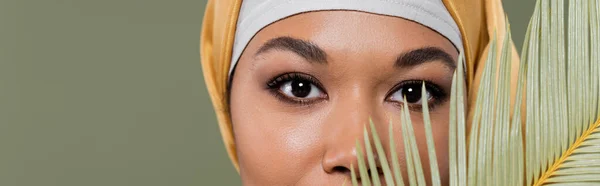 Обрезанный вид мусульманской многорасовой женщины с макияжем глядя на камеру вблизи экзотического листа изолированы на зеленый, баннер — стоковое фото