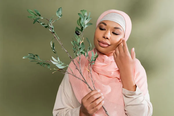 Позитивная многорасовая женщина в розовом хиджабе держит оливковую ветвь и трогает идеальное лицо на зеленом фоне — стоковое фото