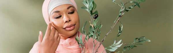 Mujer musulmana multirracial complacida con rama de olivo tocando la cara perfecta sobre fondo verde, pancarta — Stock Photo