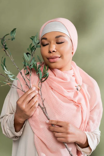 Joyful multiracial woman in pink hijab holding olive branch while posing with closed eyes on green background — Stock Photo