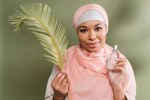 Young multiracial woman in pink hijab holding cosmetic serum and exotic leaf while looking at camera on green background — Stock Photo