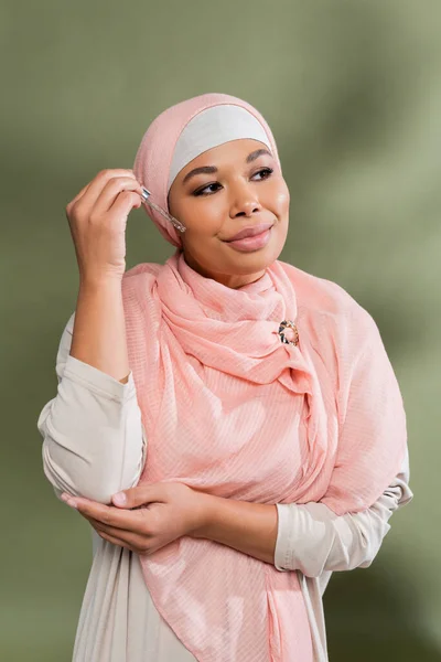 Erfreut multirassische Frau im rosa muslimischen Hijab Anwendung kosmetischen Serums auf grünem Hintergrund — Stock Photo