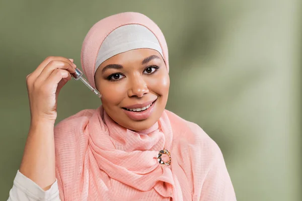 Joyful multiracial muslim woman in pink hijab applying moisturizing serum on green background — Stock Photo