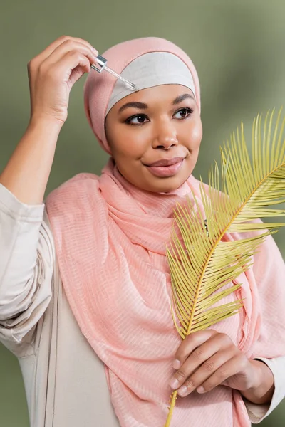 Mujer musulmana multirracial en hiyab rosa sosteniendo hoja de planta exótica y aplicando suero hidratante sobre fondo verde - foto de stock