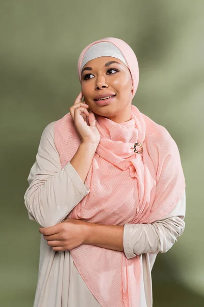 Mujer multirracial con estilo en abaya vestido y rosa hijab tocando la cara y mirando hacia otro lado sobre fondo verde - foto de stock
