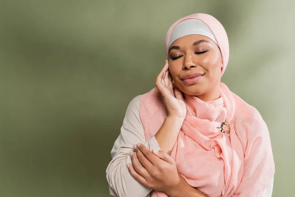 Mujer multirracial feliz en hijab rosa posando con los ojos cerrados y tocando la cara perfecta sobre fondo verde - foto de stock