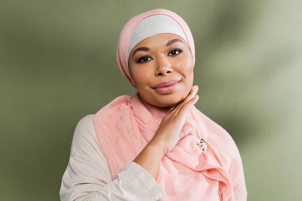 Довольная многорасовая мусульманка в розовом хиджабе трогает идеальное лицо и смотрит в камеру на зеленом фоне — стоковое фото