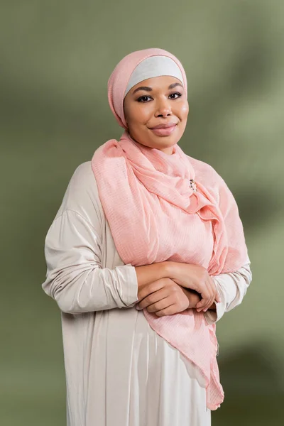 Mujer multirracial feliz en hijab rosa y vestido abaya tradicional sonriendo a la cámara sobre fondo verde - foto de stock