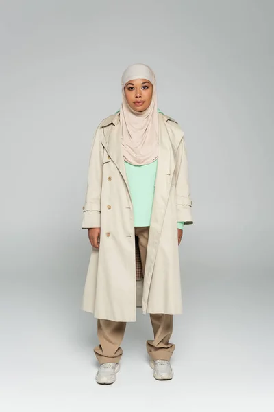 Longitud completa de la mujer multirracial en hijab y gabardina con zapatillas de deporte mirando a la cámara mientras está de pie sobre fondo gris - foto de stock