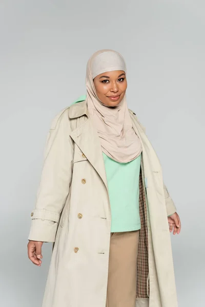 Positive multiracial model in hijab and trendy trench coat smiling at camera isolated on grey — Stock Photo
