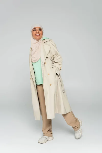 Full length of multiracial muslim woman in hijab and trench coat with sneakers posing with hand in pocket on grey background — Stock Photo