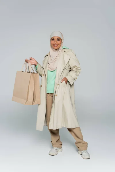 Повна довжина багаторасової жінки в хіджабі тримає сумки для покупок, позуючи рукою в кишені траншеї пальто на сірому — стокове фото