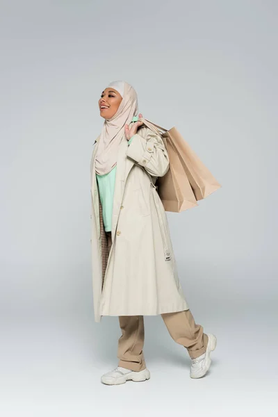 Full length of happy multiracial woman in hijab and stylish casual attire walking with shopping bags on grey background — Stock Photo