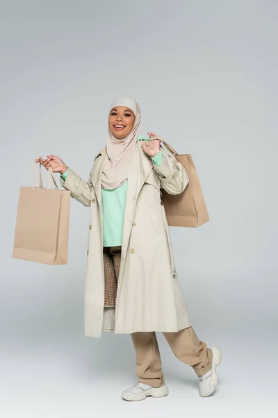Повна довжина мусульманської багаторасової жінки в траншеї пальто і хіджаб, що стоїть з сумками для покупок на сірому фоні — Stock Photo