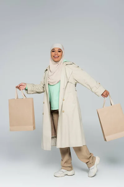 Pleine longueur de femme multiraciale heureuse dans des vêtements décontractés élégants et hijab posant avec des sacs à provisions sur fond gris — Photo de stock