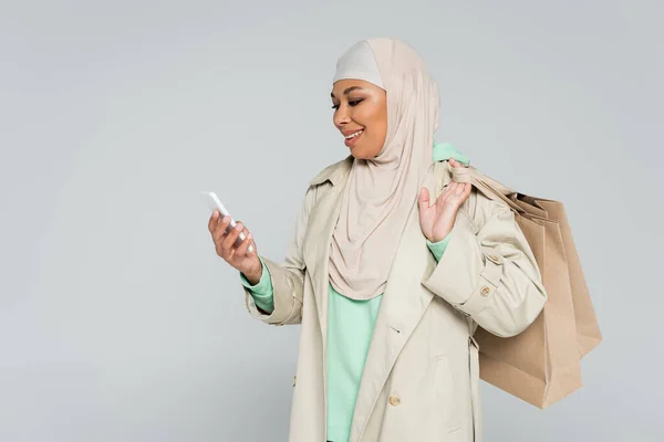 Усміхнена мультирасова жінка в траншеї пальто і хіджаб тримає сумки і дивиться на смартфон ізольовано на сірому — стокове фото