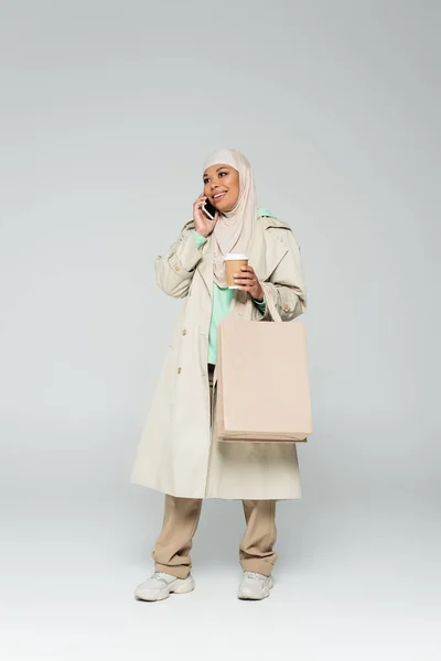 Full length of joyful multiracial muslim woman with coffee to go and shopping bags talking on smartphone on grey — Stock Photo