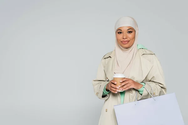 Mujer multirracial de moda en hijab y gabardina que sostiene la bebida para llevar y el bolso de compras aislado en gris - foto de stock