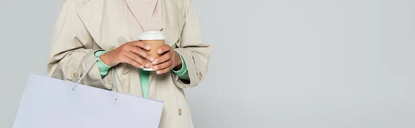 Частковий вигляд багаторасової жінки в траншеї пальто тримає сумку для покупок і одноразову чашку ізольовано на сірому, банер — стокове фото