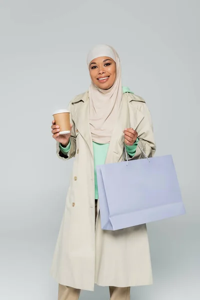 Cheerful multiracial woman in hijab and trench coat posing with paper cup and shopping bag isolated on grey — Stock Photo