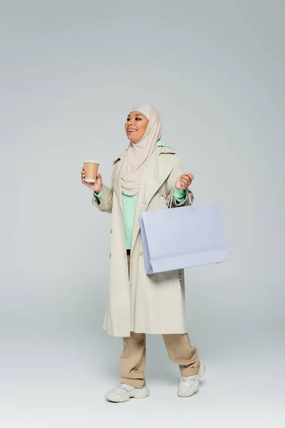 Full length of carefree multiracial woman in trendy outfit and hijab holding shopping bag and takeaway drink on grey — Stock Photo