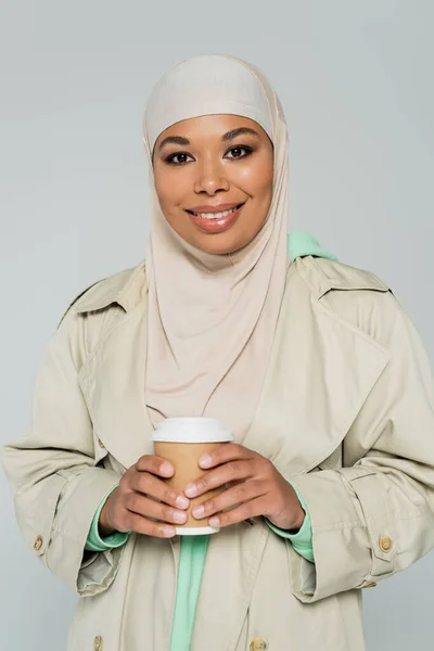 Joven mujer multirracial en hijab y elegante gabardina sosteniendo bebida para llevar y sonriendo a la cámara aislada en gris - foto de stock
