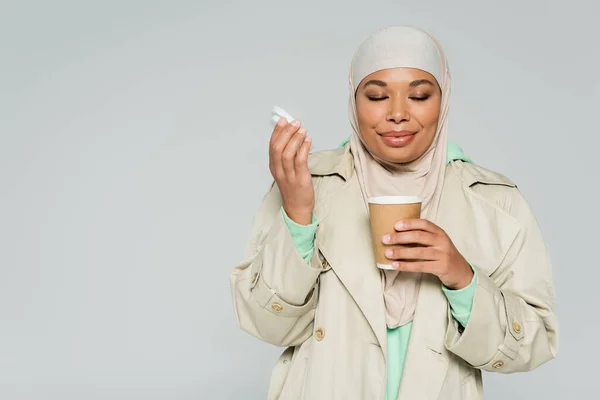 Young multiracial woman in trench coat and hijab enjoying flavor of aromatic coffee in paper cup isolated on grey — Stock Photo