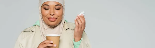 Mujer musulmana multirracial complacido con los ojos cerrados disfrutando del sabor del café en taza de papel aislado en gris, pancarta - foto de stock