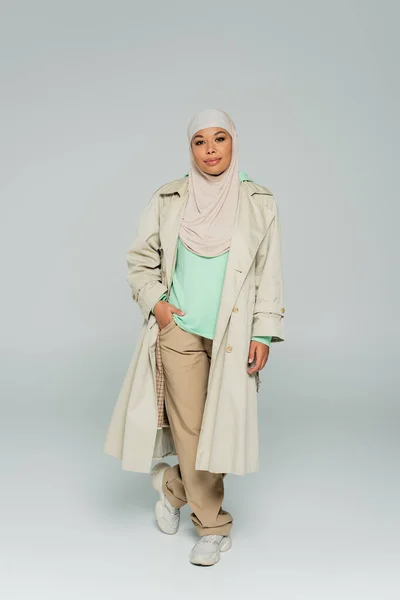 Full length of multiracial woman in fashionable casual clothes and hijab standing with hand in pocket on grey background — Stock Photo