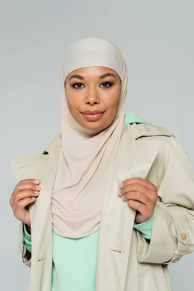 Retrato de mujer multirracial con estilo en hijab y gabardina mirando a la cámara aislada en gris - foto de stock