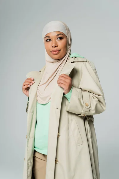 Mujer multirracial de moda en hijab y elegante gabardina mirando hacia otro lado y sonriendo aislado en gris - foto de stock