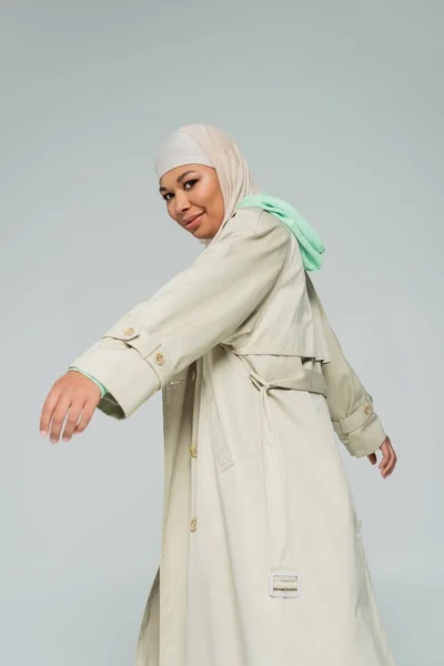 Joven y despreocupada mujer multirracial en hijab y gabardina sonriendo a la cámara mientras posando aislada en gris - foto de stock