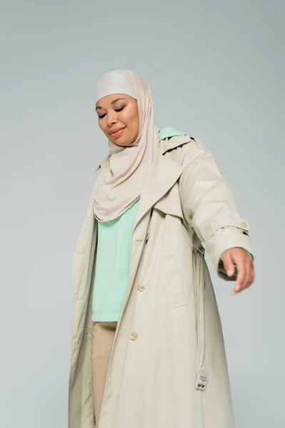 Joven y sonriente mujer multirracial en hijab y elegante gabardina posando aislado en gris - foto de stock