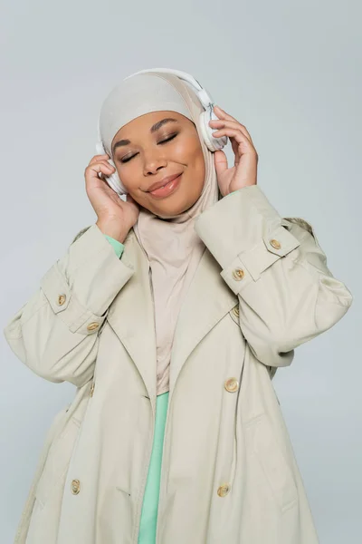 Радостная и стильная многорасовая мусульманка с закрытыми глазами слушающая музыку в беспроводных наушниках, изолированных на сером — стоковое фото