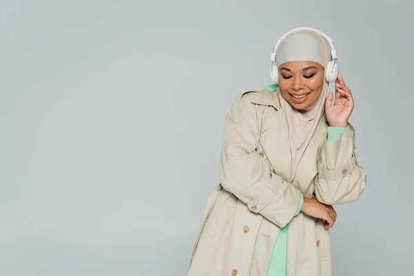 Überglückliche multirassische Frau in Trenchcoat und Hijab, die Musik in drahtlosen Kopfhörern isoliert auf grau hört — Stockfoto
