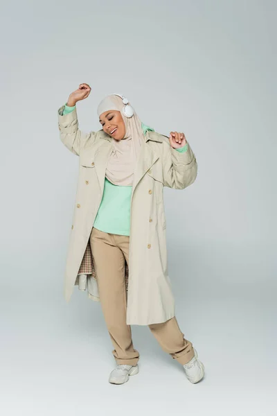 Full length of carefree and fashionable multiracial muslim woman listening music in headphones and dancing on grey — Stock Photo