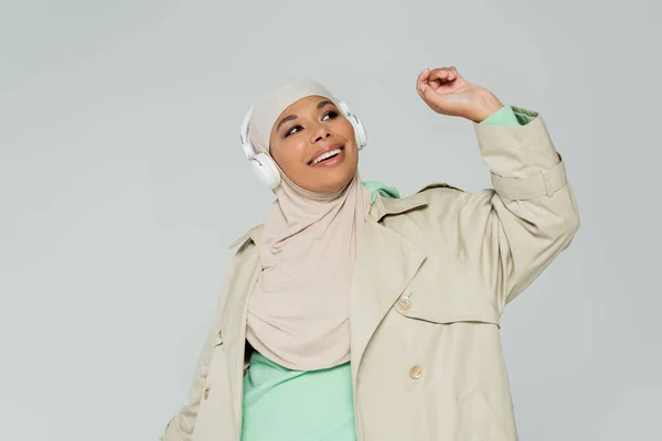 Overjoyed multiracial woman in trench coat and hijab listening music in wireless headphones isolated on grey — Stock Photo