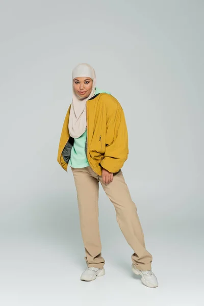 Full length of multiracial woman in hijab and trendy casual outfit standing and smiling at camera on grey background — Stock Photo