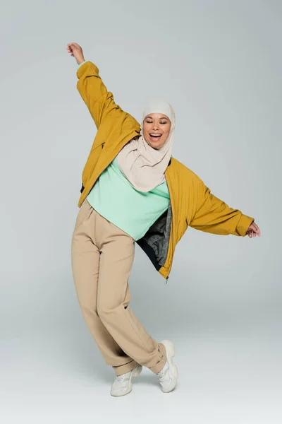 Полная длина многорасовых мусульманских женщин в желтой куртке бомбардировщика и бежевых брюках позирует с протянутыми руками на сером фоне — стоковое фото