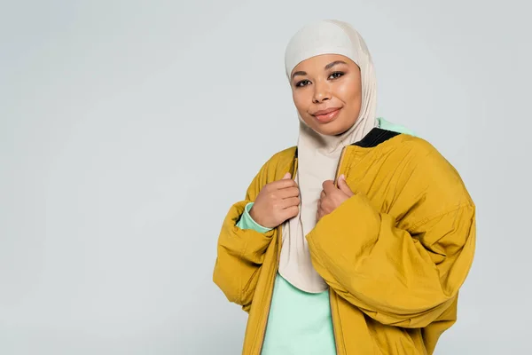 Musulmana mujer multirracial sonriendo a la cámara mientras posando en hijab y chaqueta bombardero amarillo aislado en gris - foto de stock