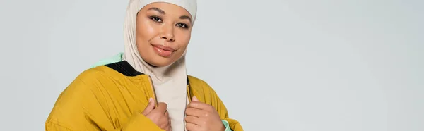 Alegre mujer musulmana multirracial en hijab y chaqueta con estilo amarillo mirando la cámara aislada en gris, pancarta - foto de stock