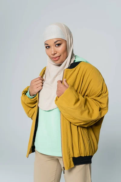 Молода і щаслива багаторасова жінка в жовтій куртці бомбардувальника і хіджаб посміхається на камеру ізольовано на сірому — стокове фото