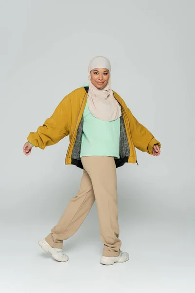 Повна довжина веселої мультирасової мусульманської жінки в жовтій куртці бомбардувальника і бежевих штанах, що ходять на сірому фоні — стокове фото
