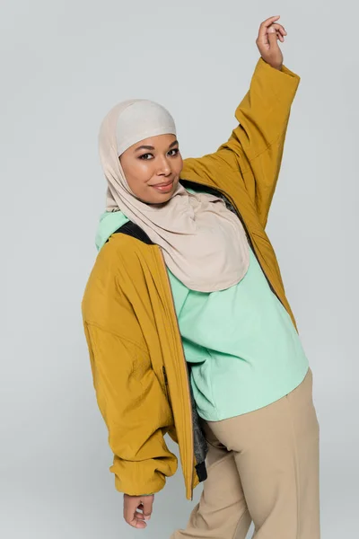 Femme multiraciale à la mode en veste de bombardier jaune et hijab posant avec la main levée isolé sur gris — Photo de stock