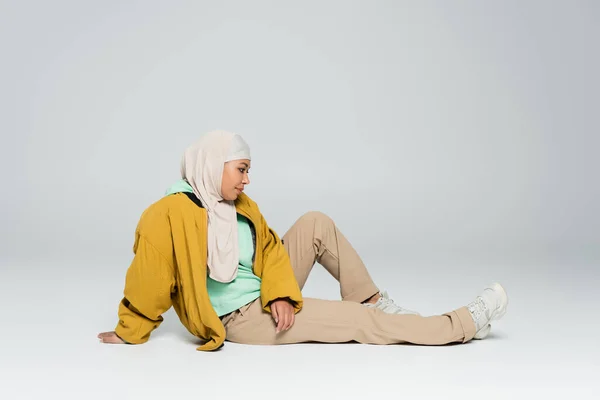 Longitud completa de la mujer multirracial en hijab y chaqueta amarilla con pantalones beige sentado sobre fondo gris - foto de stock