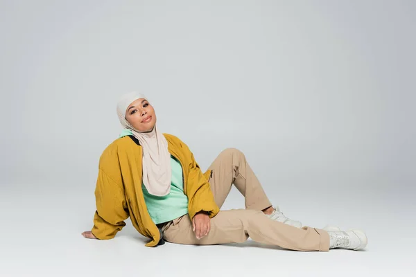 Full length of carefree multiracial woman in hijab and fashionable casual attire sitting on grey background — Stock Photo
