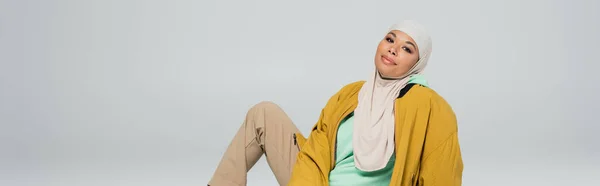 Donna musulmana multirazziale alla moda in hijab e giacca gialla sorridente alla macchina fotografica mentre seduto isolato sul grigio, banner — Foto stock