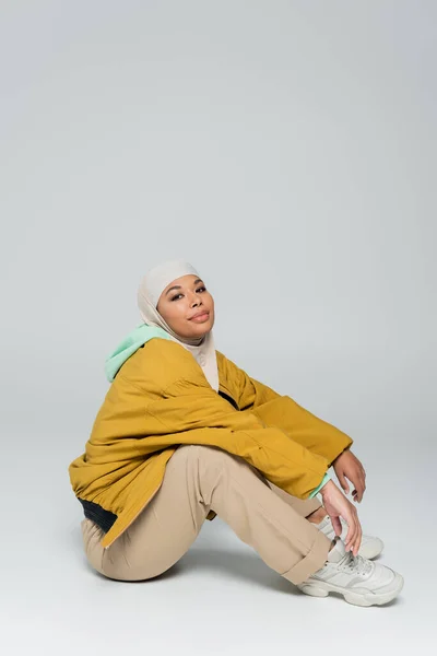 Longitud completa de la mujer musulmana multirracial complacida sentada en pantalones beige y chaqueta amarilla sobre fondo gris - foto de stock